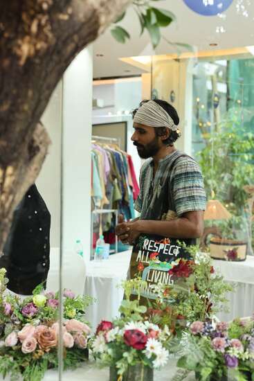 A man with a headband stands indoors, surrounded by vibrant floral arrangements. He carries a colorful tote bag reading, “RESPECT ALL LIVING THINGS,” and wears a striped shirt.