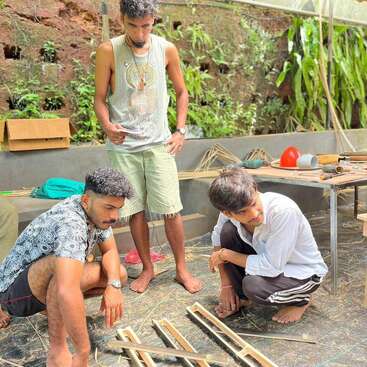 The image depicts three men working on a project, surrounded by various materials and tools, with a focus on crafting wooden frames.