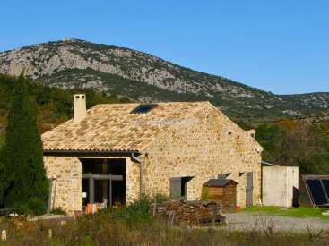 L'image représente une maison rustique en pierre avec un toit en pente, située dans une zone rurale avec un arrière-plan montagneux et un ciel bleu clair.