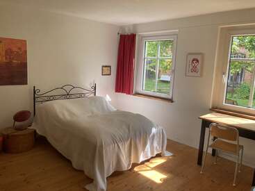 A bright, minimalist bedroom with a simple metal-frame bed, white bedding, wooden floor, red curtain, desk, chair, two windows, and light streaming in.