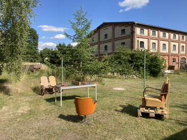 A grassy yard features eclectic outdoor furniture around a white table, with an old brick building, trees, and blue sky with fluffy clouds in the background.