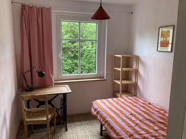 A small, cozy bedroom with a single bed, striped orange bedding, wooden desk, chair, lamp, shelves, red curtain, window, framed artwork, and a pendant light.