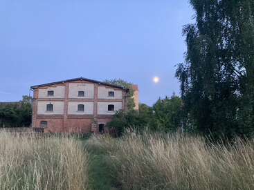 An old, abandoned brick building stands amid tall grass at dusk, with a full moon rising in a clear, pale blue evening sky. Trees surround.
