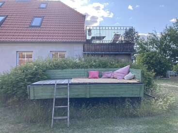 A cozy outdoor daybed with colorful pillows sits on a raised wooden platform, surrounded by greenery, in front of a house with a red-tiled roof.