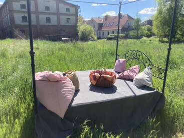 A cozy outdoor bed with pillows and baskets sits in a grassy field, surrounded by green nature and houses, beneath a sunny blue sky with clouds.
