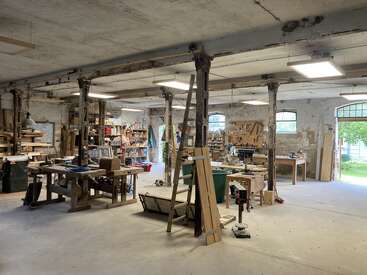 This image shows a spacious, well-lit woodworking shop with workbenches, tools, lumber, shelves, rustic columns, concrete floors, and large windows, giving an industrious, organized atmosphere.