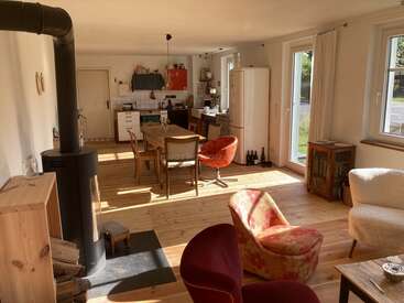 Sunlit open living space with wooden floors, cozy seating, a wood-burning stove, dining table, and an inviting kitchen. Large windows provide ample daylight and garden view.