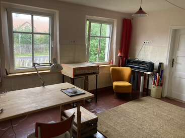 A cozy workspace featuring wooden desks, a yellow armchair, large windows, red curtains, a desk lamp, rolled-up posters, a printer, and simple decor elements.