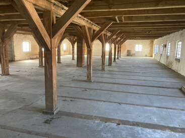 The image depicts a spacious, empty warehouse interior with exposed wooden beams, concrete floor, large windows, natural light, and rustic brick walls, creating an industrial, historic atmosphere.