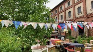 A festive outdoor scene features colorful bunting, tables, and chairs arranged in a garden near a brick building. Trees and greenery add to the cheerful atmosphere.