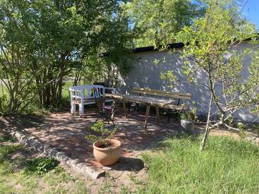 A cozy outdoor patio with a wooden table, benches, and a white garden bench, surrounded by greenery, trees, potted plants, and a sunlit brick floor.