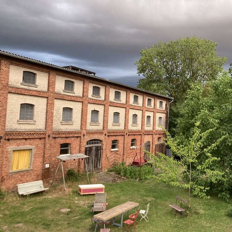 An old brick building with many windows stands beside a yard with benches and tables. Tall green trees and dramatic, dark clouds fill the background.