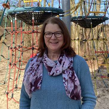 A smiling woman wearing glasses, a blue sweater, and a floral scarf stands in front of a colorful rope playground structure on a sunny day outside.