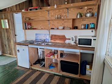 Cette cuisine rustique et confortable comprend des étagères ouvertes en bois, un double évier, un petit réfrigérateur, un micro-ondes, une bouilloire, de la vaisselle, des ustensiles de cuisine et divers articles de cuisine, le tout éclairé par la lumière naturelle.