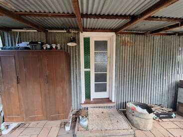 Une cabane rustique en tôle ondulée, usée par les intempéries, avec une porte vitrée blanche, une armoire brune, un vieux tapis, une baignoire en métal, des objets éparpillés et une toiture en bois apparente au-dessus.