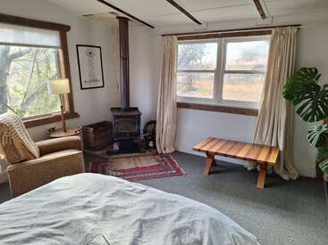 Cette chambre confortable comprend un lit, un fauteuil, un poêle à bois, une table rustique, de grandes fenêtres avec des rideaux, une plante feuillue, des œuvres d'art encadrées et une atmosphère chaleureuse et accueillante.