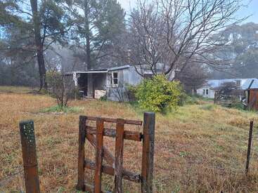 Un vieux portail en bois mène à une maison abandonnée, entourée d'herbe envahissante, d'arbres dénudés et de brume, évoquant un sentiment d'isolement tranquille et de décadence rustique.