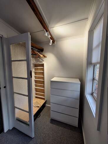 Ce petit placard lumineux est composé d'une commode blanche, d'une penderie ouverte avec des cintres, d'un sol recouvert de moquette, d'une porte en verre dépoli, d'une fenêtre avec des stores et d'une poutre en bois apparente au plafond.