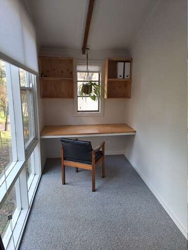 Cette image montre un petit bureau minimaliste avec un bureau en bois, une chaise, une plante suspendue, des étagères, de grandes fenêtres, de la moquette et de la lumière naturelle.