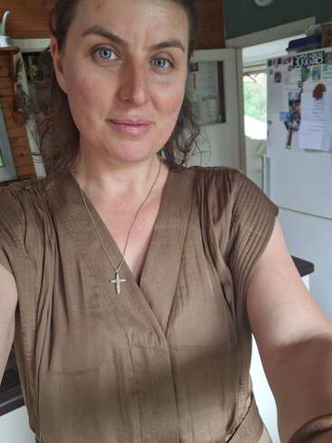 Une femme aux yeux bleus et aux cheveux bruns ondulés prend un selfie dans une cuisine. Elle porte une robe brune et un collier en forme de croix en argent.