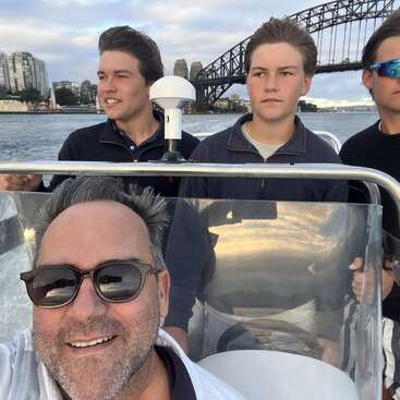 Four people are on a boat, smiling and enjoying a ride near the Sydney Harbour Bridge, with city buildings and water in the background under a cloudy sky.