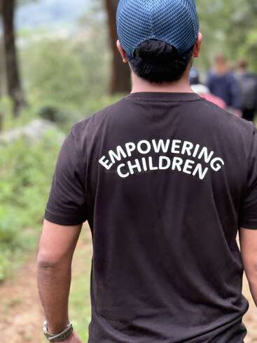 A person wearing a blue cap and a black T-shirt with the words "EMPOWERING CHILDREN" walks outdoors along a forested path, surrounded by greenery.