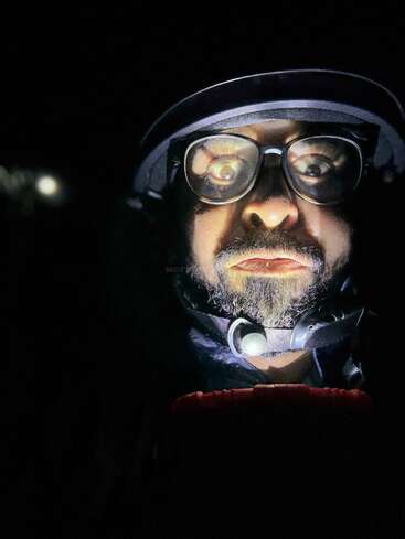 A man wearing glasses and a helmet is illuminated from below, creating a dramatic, eerie effect. His wide eyes and expression appear intense in darkness.