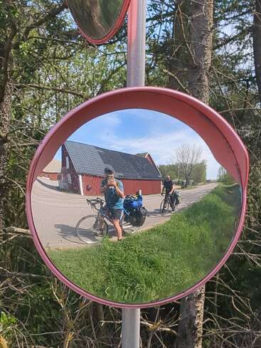 Ein runder Verkehrsspiegel spiegelt zwei Radfahrer mit beladenen Fahrrädern auf einer Landstraße neben einem roten Haus, umgeben von Grün und Bäumen, unter einem sonnigen Himmel.
