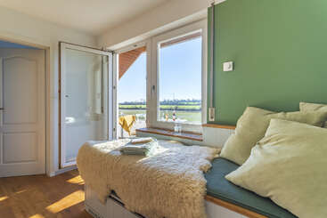 Ein gemütliches, sonnendurchflutetes Zimmer mit einer gepolsterten Bank mit flauschigen Überwürfen, großen Fenstern, einer grünen Akzentwand und einer offenen Tür mit Blick auf eine malerische Landschaft.