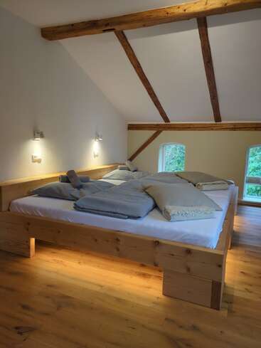 Esta acogedora habitación abuhardillada cuenta con una gran cama de madera, iluminación tenue, vigas de madera vistas, suelos de madera, ropa de cama fresca y dos ventanas que ofrecen una vista verde y serena.