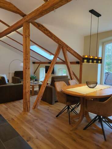 Esta imagen muestra un acogedor y moderno loft con vigas de madera vistas, una mesa de comedor redonda, cuatro sillas, iluminación colgante, sofás marrones, suelos de madera y grandes ventanales.