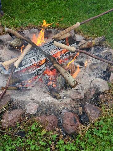 Una acogedora hoguera rodeada de piedras sobre hierba verde. Varios palos con masa de pan se asan sobre las llamas, creando una escena al aire libre cálida y acogedora.