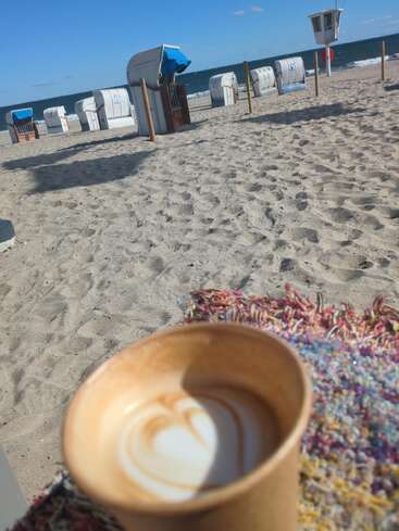 Una taza de café con leche en forma de corazón sobre una manta de colores en una playa de arena, con sillas de playa y el océano de fondo.