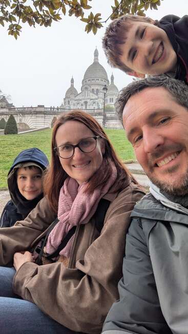 Une famille de quatre personnes, tout sourire, prend un selfie en plein air devant la basilique du Sacré-Cœur, au milieu de la verdure, des arbres et d'un ciel couvert.
