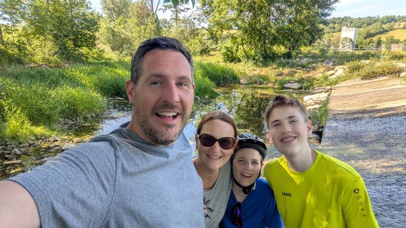 Une joyeuse famille de quatre personnes pose pour un selfie en plein air, au bord d'un ruisseau pittoresque et verdoyant. Tout le monde sourit, profitant ensemble d'une belle journée ensoleillée.