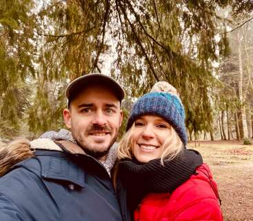 Un couple heureux, chaudement vêtu de vestes et de chapeaux, sourit ensemble en plein air dans une zone boisée, entouré de grands arbres verts sous un ciel couvert.