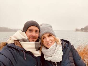 Un couple vêtu de manteaux d'hiver, d'écharpes et de bonnets se tient tout près l'un de l'autre, souriant au bord d'un lac calme par une journée nuageuse, entouré de roseaux bruns et d'arbres.