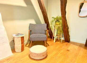 Un coin confortable comprend un fauteuil à motifs, une petite table d'appoint en bois avec des magazines, un pouf rond, une plante feuillue sur un support, des poutres rustiques et un miroir mural.