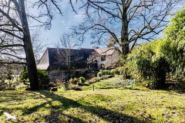 Une charmante maison en pierre nichée parmi de grands arbres et une végétation luxuriante, baignée de soleil, avec des ombres qui s'étendent sur la pelouse paisible et légèrement envahie par la végétation.