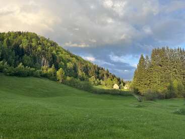 A peaceful green meadow stretches under a dramatic sky. Sunlight touches the forested hills and a few houses nestled quietly in the distance, creating a tranquil scene.