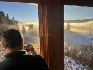 A man stands inside a wooden cabin, photographing a breathtaking winter landscape outside the window, filled with mist, pine trees, snow, and golden sunlight.