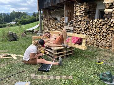 Two people build a rustic outdoor bench from wooden pallets. One lounges on the bench while the other uses a drill, surrounded by stacked firewood and green countryside.