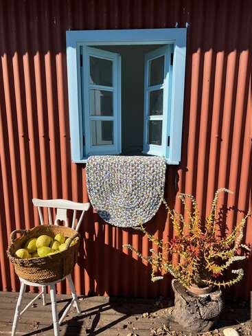 Una escena rústica en la que se aprecia una ventana abierta con marco azul, una pared roja con textura, una silla blanca con una cesta de limones, una alfombra de colores y una planta en maceta de vivos colores.