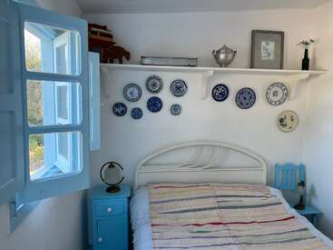 Un acogedor dormitorio con toques de color azul claro, una ventana abierta, paredes blancas, platos decorativos azules y blancos colgados en la pared, una estantería con libros y muebles de estilo rústico.