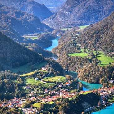 A picturesque village nestles among lush green hills, winding turquoise river flows through the valley, surrounded by forested mountains under a clear, blue sky. Peaceful paradise.