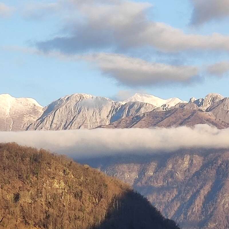 Das Bild zeigt eine majestätische Bergkette mit schneebedeckten Gipfeln vor einem blauen Himmel mit Wolken und einem Tal mit Bäumen und einer tief hängenden Wolke.