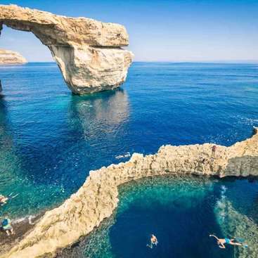 The image depicts a serene natural setting with a large rock formation and a pool of water, featuring people swimming and snorkeling in the crystal-clear blue waters.
