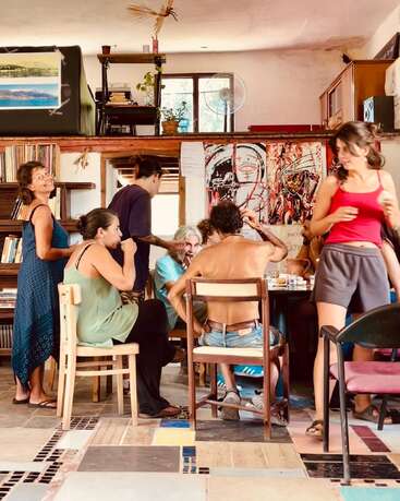 Un groupe de personnes se réunit dans une pièce confortable et artistique, engageant la conversation et partageant un repas. L'espace est rempli de livres, d'œuvres d'art et d'une énergie vibrante.