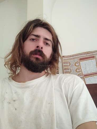 The image depicts a man with long brown hair and a white t-shirt, standing in front of a white wall with a painting on it.