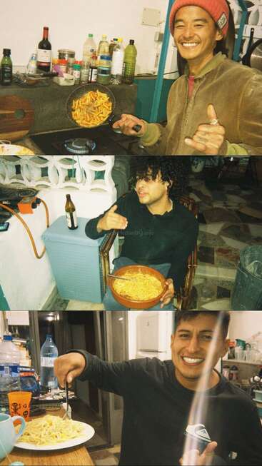 Tres amigos disfrutan juntos de la pasta en una acogedora cocina, sonriendo y haciendo señas con las manos, con varias bebidas, utensilios e ingredientes visibles a su alrededor, creando un ambiente cálido.